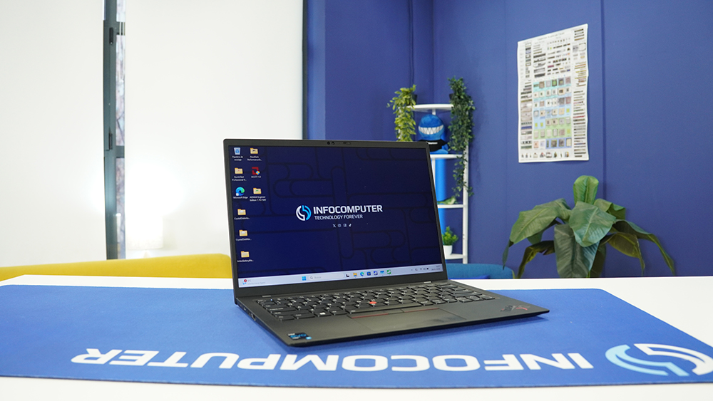 Portátil Lenovo ThinkPad X1 Carbon abierto sobre un escritorio azul de InfoComputer, mostrando el fondo de pantalla corporativo en un entorno de estudio.